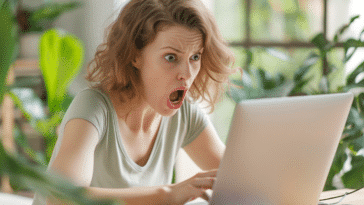 Une jeune femme assise à son bureau, devant son ordinateur portable, affiche une expression de choc avec la bouche grande ouverte et les yeux écarquillés.