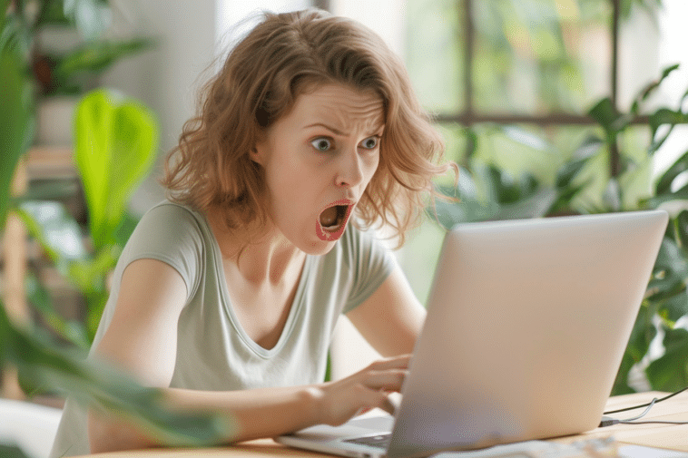 Une jeune femme assise à son bureau, devant son ordinateur portable, affiche une expression de choc avec la bouche grande ouverte et les yeux écarquillés.