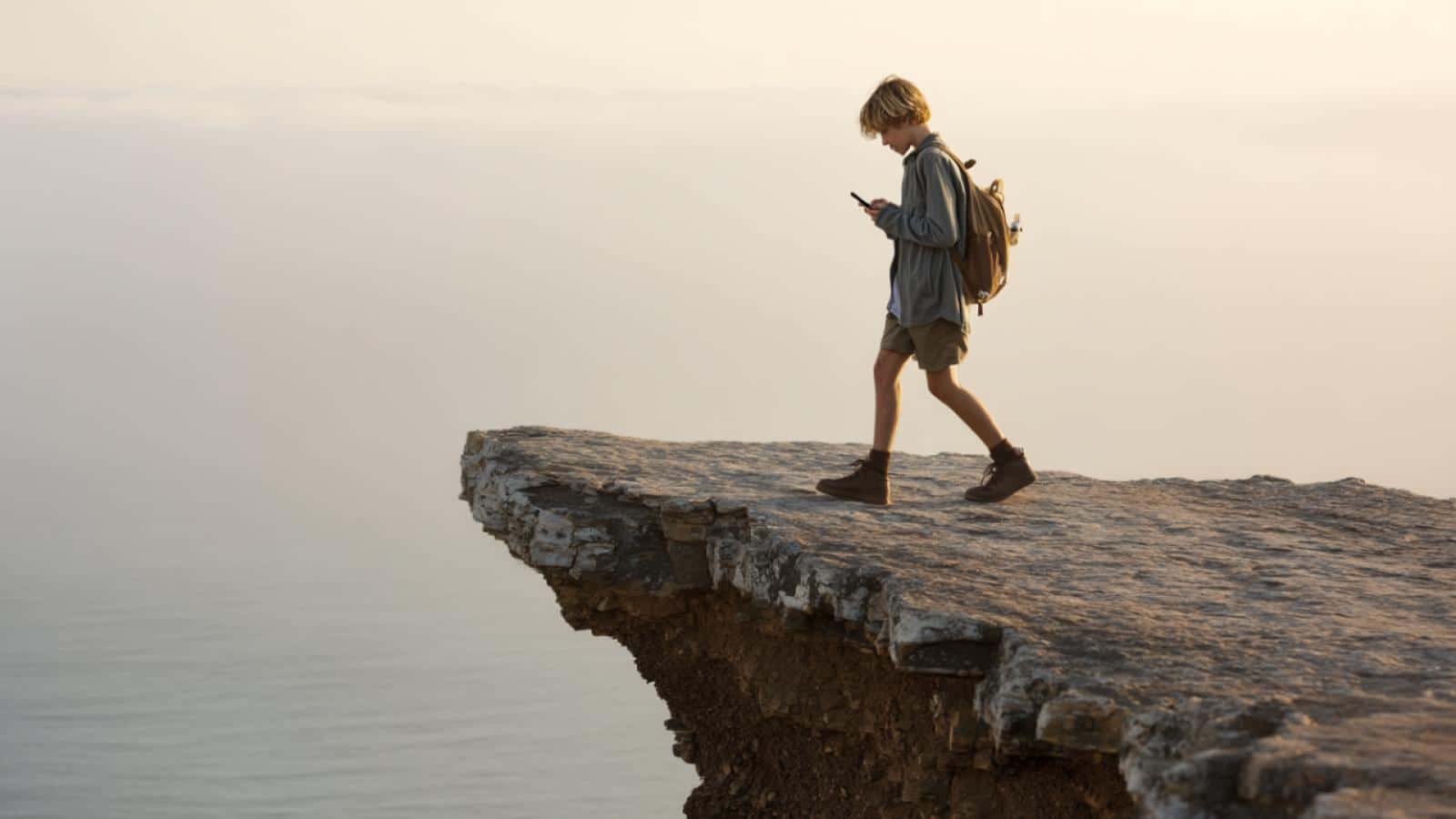 image générée d'un enfant qui discute avec un ami chatbot sur smartphone sans voir qu'il marche au bord d'une falaise
