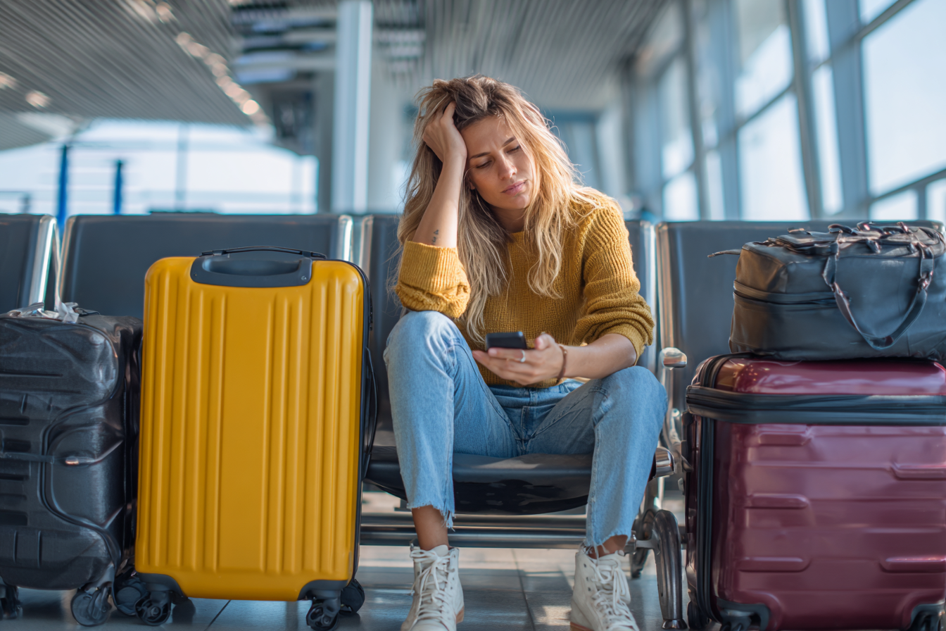 Passager frustré dans un aéroport, assis parmi ses valises, utilisant son smartphone après l’annonce d’un vol annulé.
