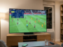 Vue de dos d'une personne assise dans un salon chaleureux, regardant un match de football entre l'OGC Nice et le RC Lens sur un grand écran plat posé sur un meuble en bois. À droite, une grande bibliothèque remplie de livres complète l'ambiance cosy
