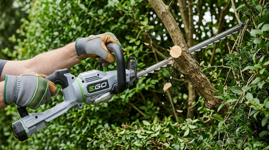 Gros plan sur la lame du taille-haie EGO Power+ HT6510E en train de couper une branche épaisse au cœur d'une haie.