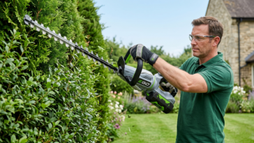 Un homme utilise le taille-haie à batterie EGO Power+ HT6510E pour entretenir une haie dense dans un jardin.