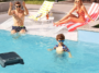 Le robot skimmer de piscine solaire Aiper EcoSurfer S2 flottant dans l'eau d'une piscine ensoleillée avec une famille en arrière-plan.