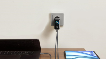 Un chargeur Anker Zolo 140W noir est branché dans une prise murale grise, alimentant simultanément un ordinateur portable et une tablette sur un bureau.