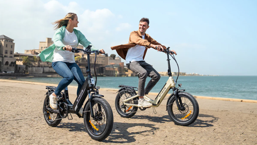 Deux personnes souriantes sur des vélos ENGWE L20 3.0 Boost roulant sur un chemin pavé au bord de la mer