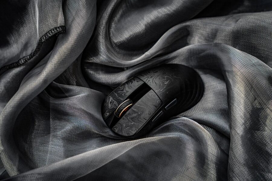 Une souris de jeu Epomaker Carbonis noire en fibre de carbone composite, posée sur un tissu soyeux et plissé de couleur gris anthracite, avec un éclairage doux.