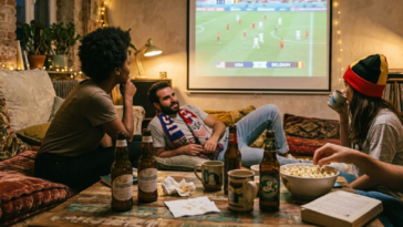 Amis regardant un match de football entre les États-Unis et la Belgique projeté sur un grand écran dans un salon chaleureux.