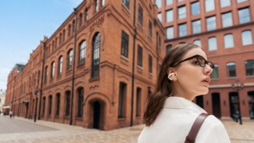 Femme portant des écouteurs Shokz OpenDots 2 blancs et des lunettes de soleil, de profil dans une rue pavée.