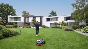 Un homme surveillant le robot tondeuse Navimow H2 tondre une vaste pelouse devant une maison moderne.