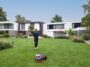 Un homme surveillant le robot tondeuse Navimow H2 tondre une vaste pelouse devant une maison moderne.