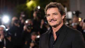 Cette photo montre Pedro Pascal, vêtu d'un costume noir élégant et d'une chemise noire, souriant devant une foule de photographes.