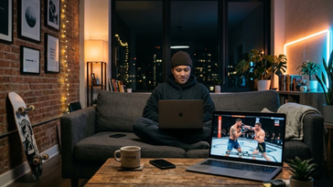 Homme assis sur un canapé dans un appartement moderne regardant un combat de MMA en direct sur son ordinateur portable.