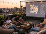 Groupe d'amis regardant le combat UFC Volkanovski vs Evloev sur écran géant depuis un rooftop au coucher du soleil